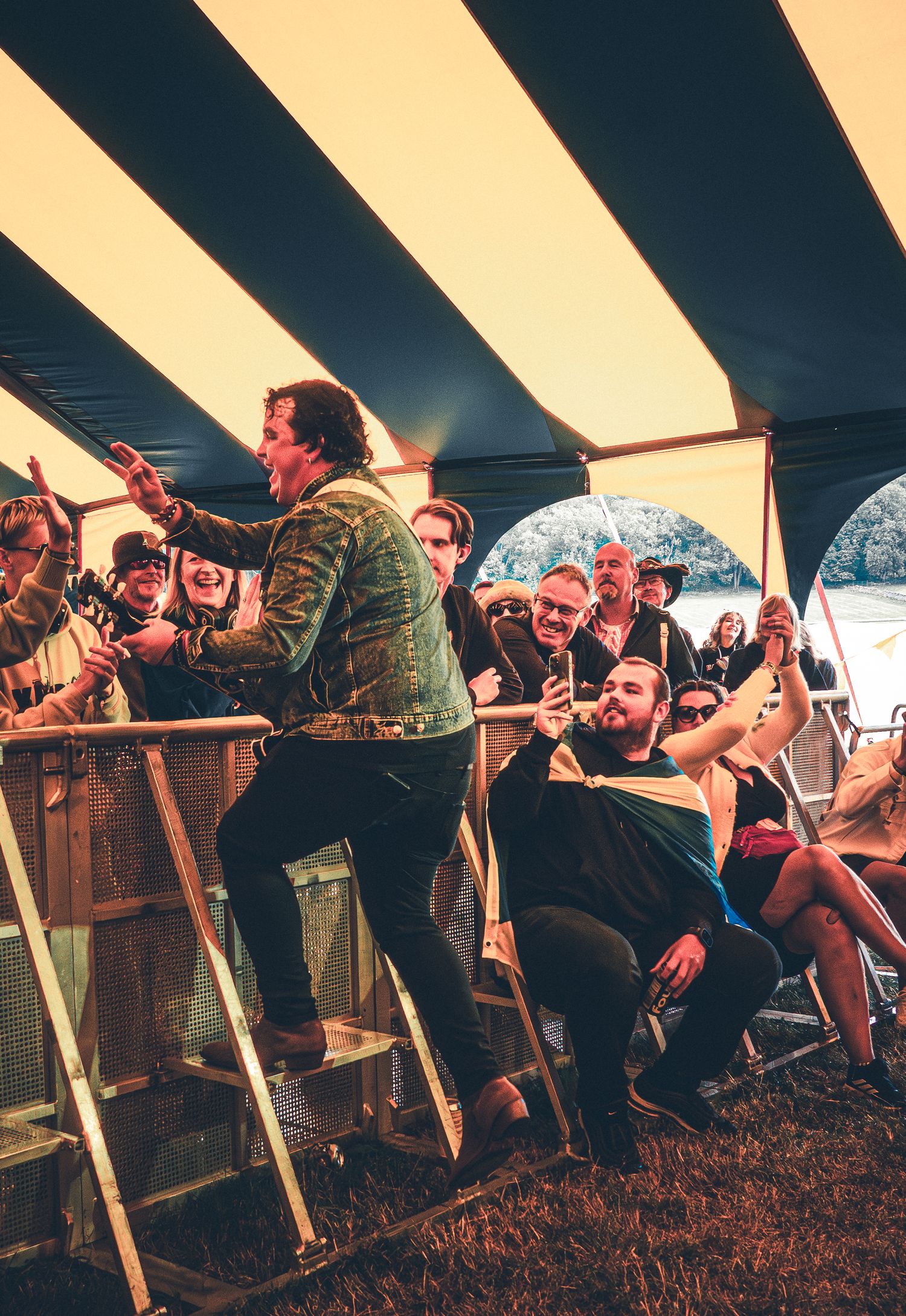 Energetic performer engages with a crowd of festival-goers under a striped tent at an outdoor music event.