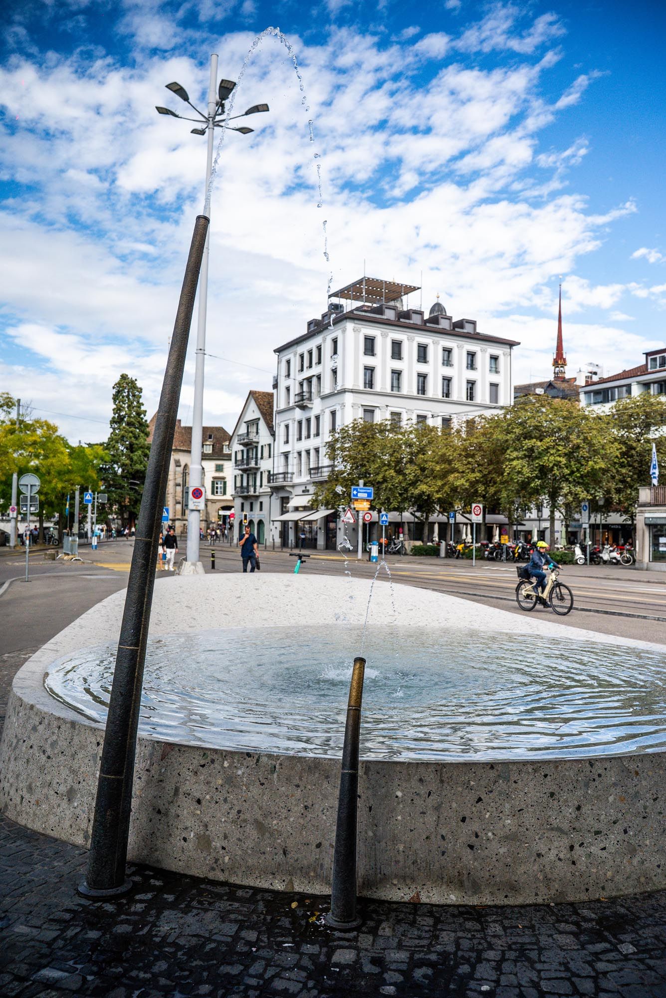 A modern fountain in a European town square with historic white buildings and blue sky with puffy clouds in the background.