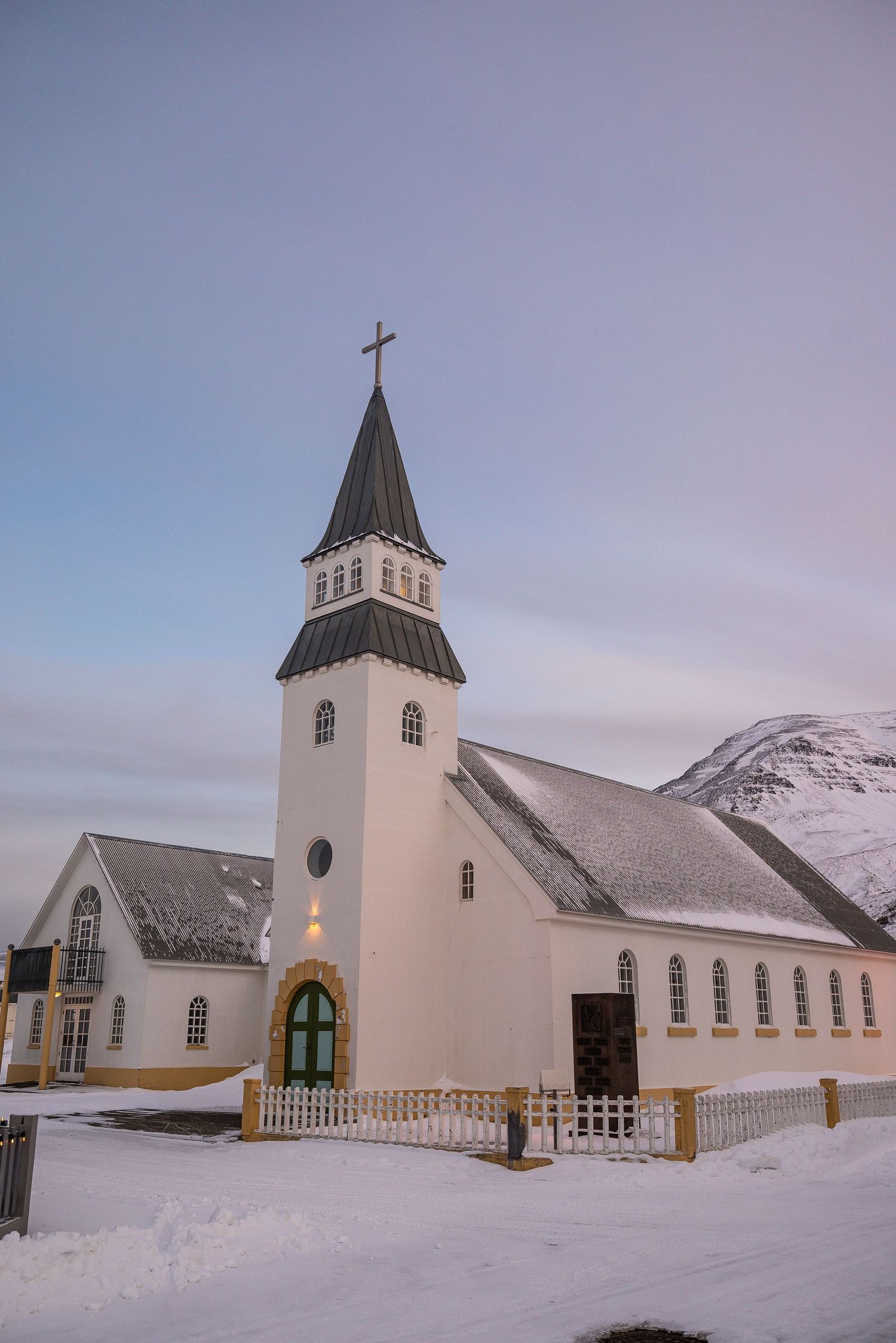 A white Nordic church with steeple stands in snowy landscape against cloudy sky and mountain backdrop in Iceland.