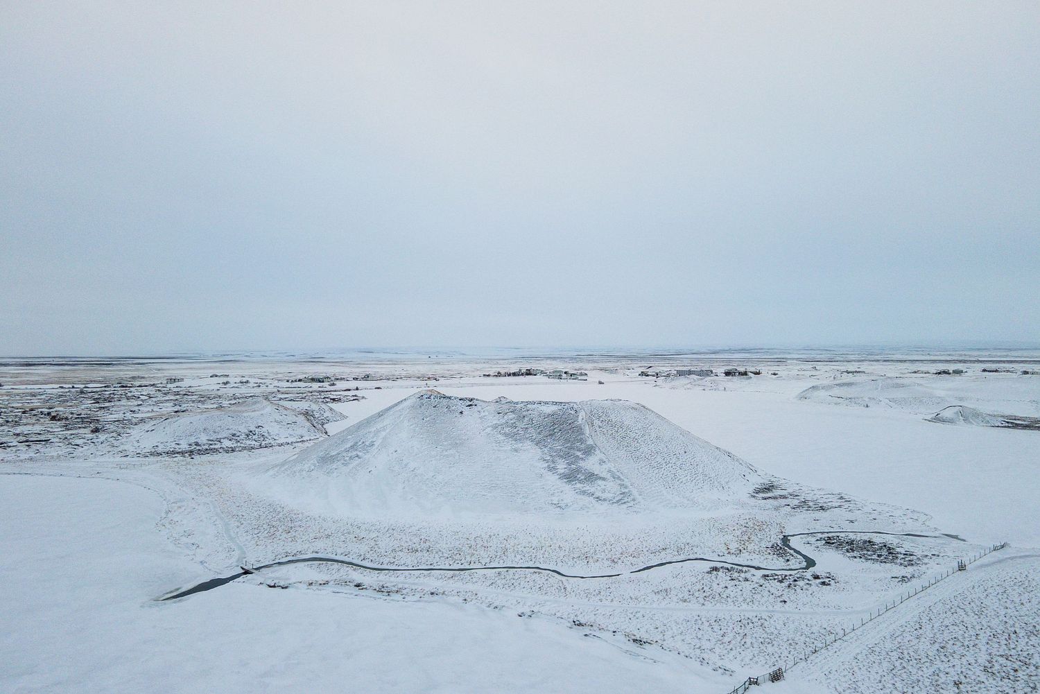 A snow-covered winter landscape with rolling hills and a stark horizon against an overcast sky.