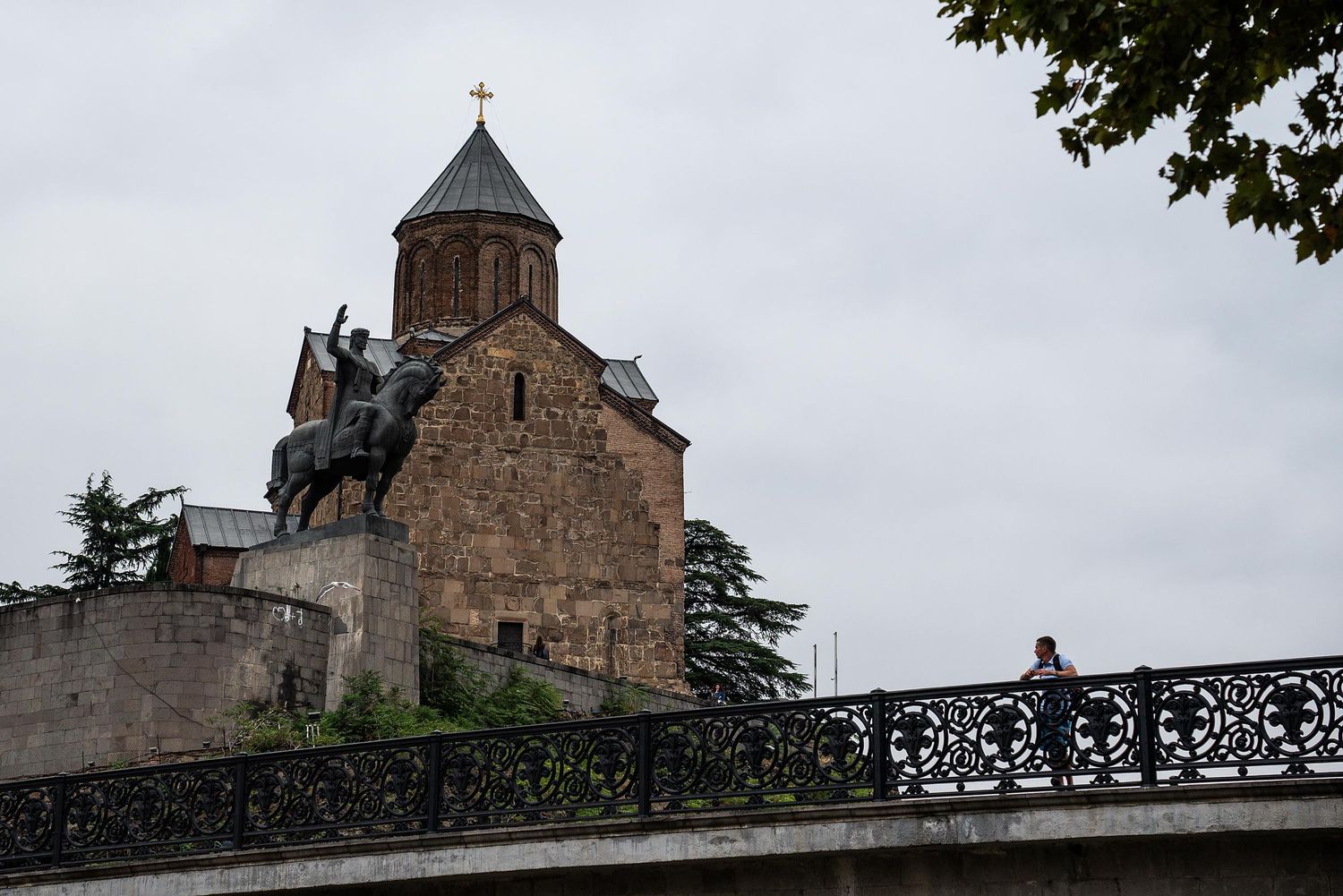 Historic Metekhi Church with equestrian statue perched on a cliff overlooking Tbilisi, Georgia against cloudy sky.