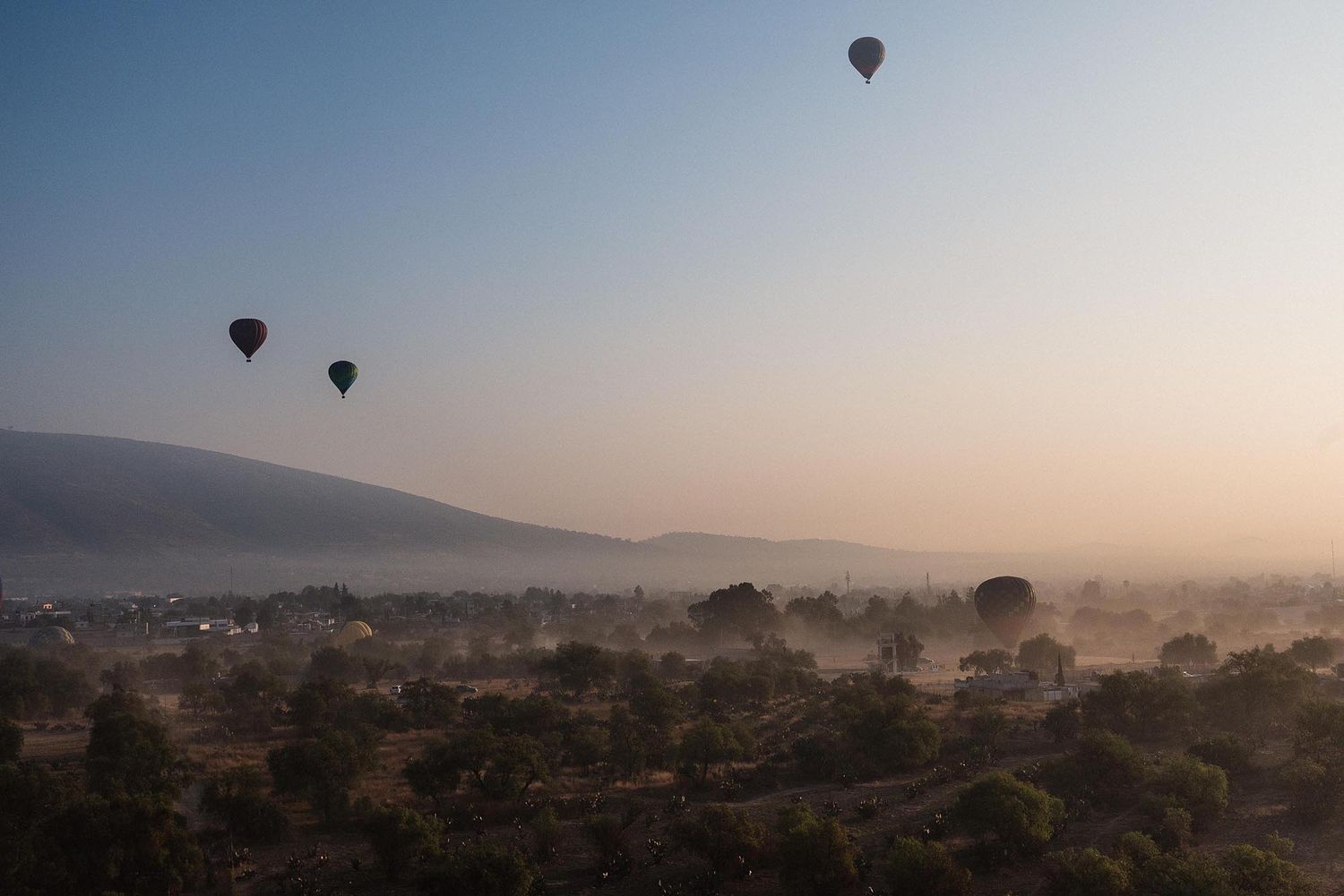 Hot air balloon floating over misty mountain landscape at sunrise with silhouetted hills and hazy atmosphere.