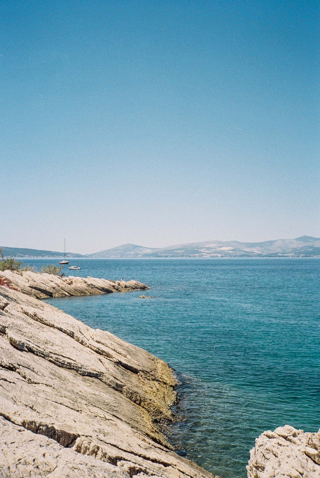 Rocky coastal outcrop with layered limestone formations jutting into bright blue Mediterranean waters on a sunny day.