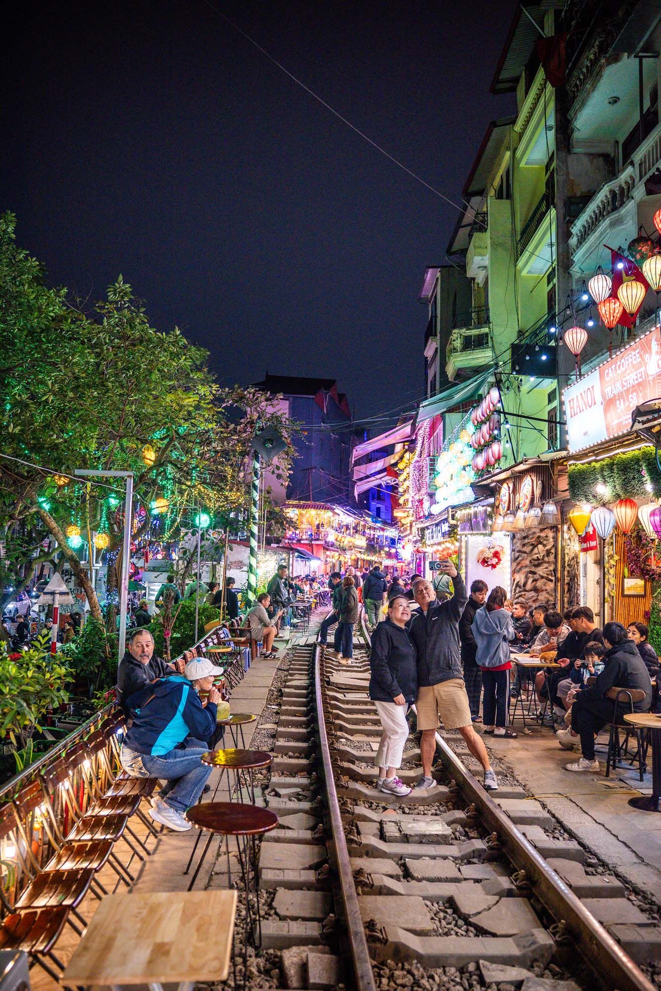 Tourists explore the vibrant Train Street in Hanoi at night with colorful lights illuminating the narrow railway tracks.