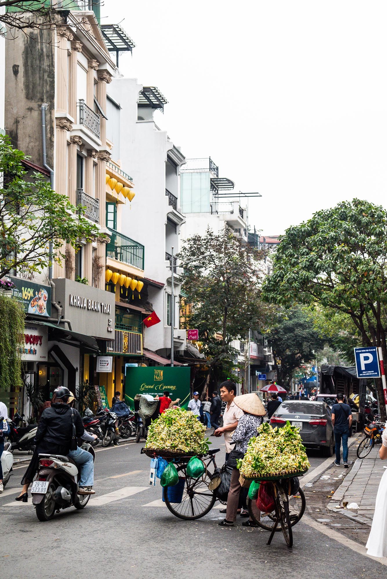 Street scene in Vietnam with flower vendors on bicycles and motorbikes traveling along a busy urban road with shops.