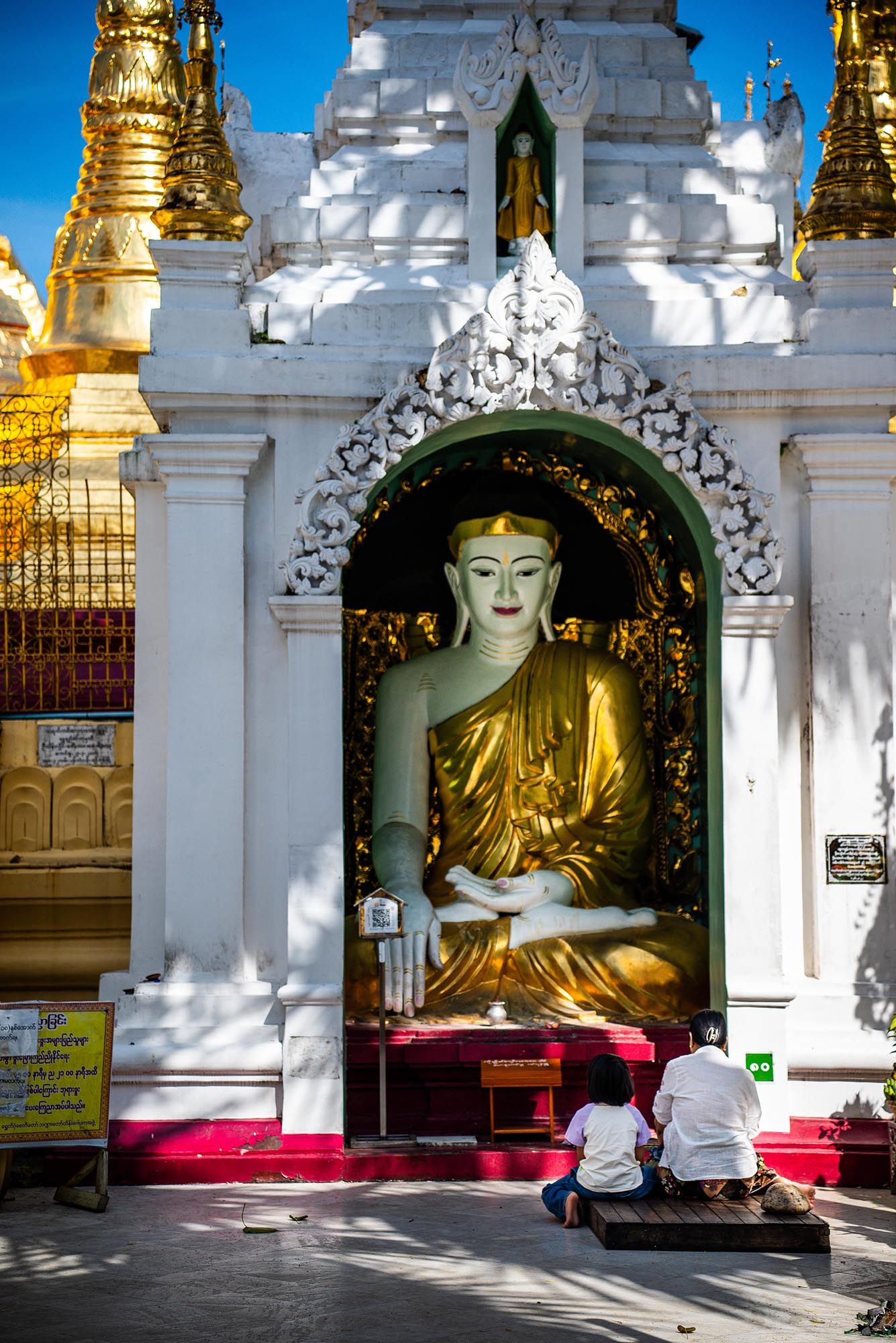 Golden Buddha statues sit in ornate white alcoves at Shwedagon Pagoda in Yangon, Myanmar, casting dramatic shadows.