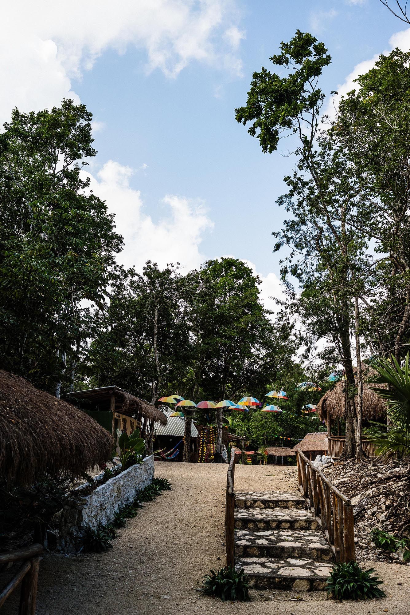 Rustic stone steps and dirt path lead to thatched-roof structures in a tropical jungle setting with blue sky overhead.