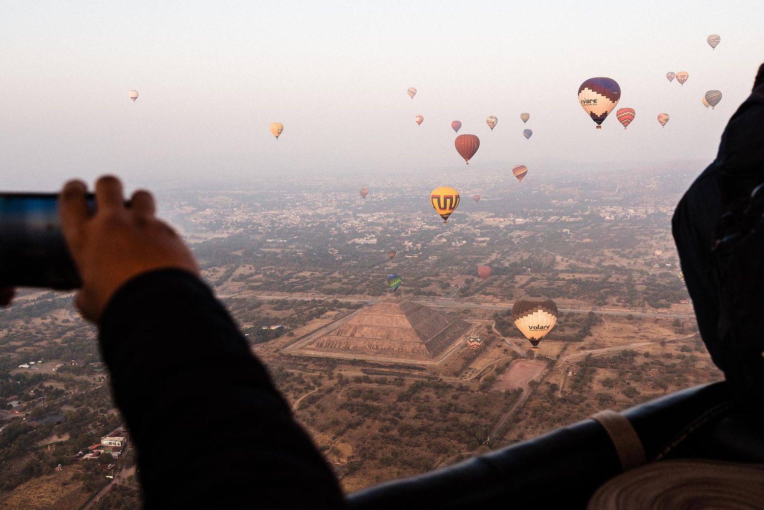 Hot air balloons float above an arid desert landscape during sunrise with silhouettes visible in the gondola baskets.