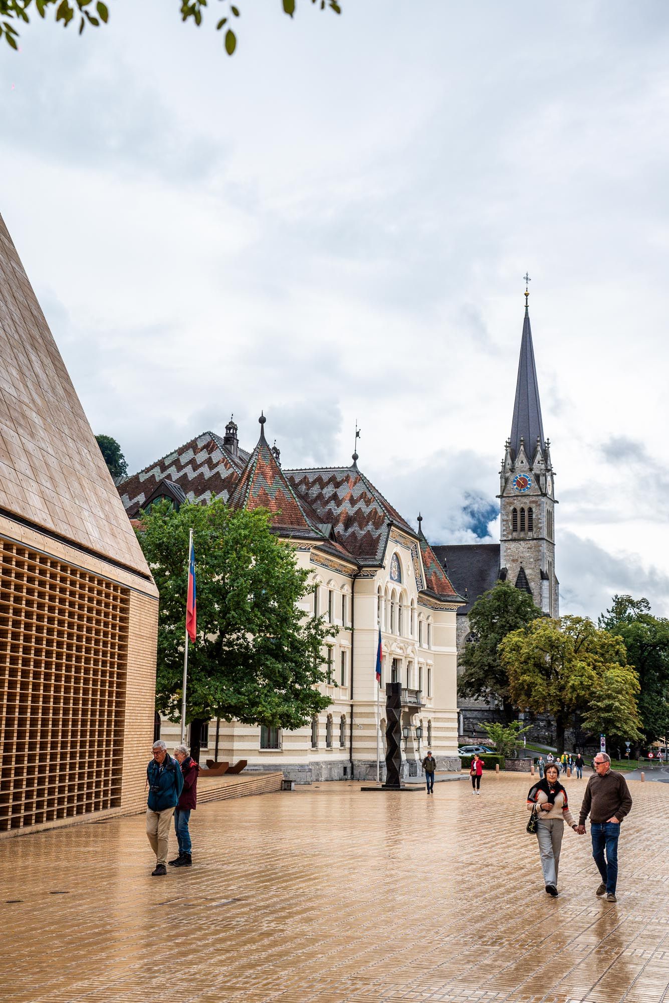 People walk along a modern plaza with brick buildings and church spires visible in the background on a cloudy day.