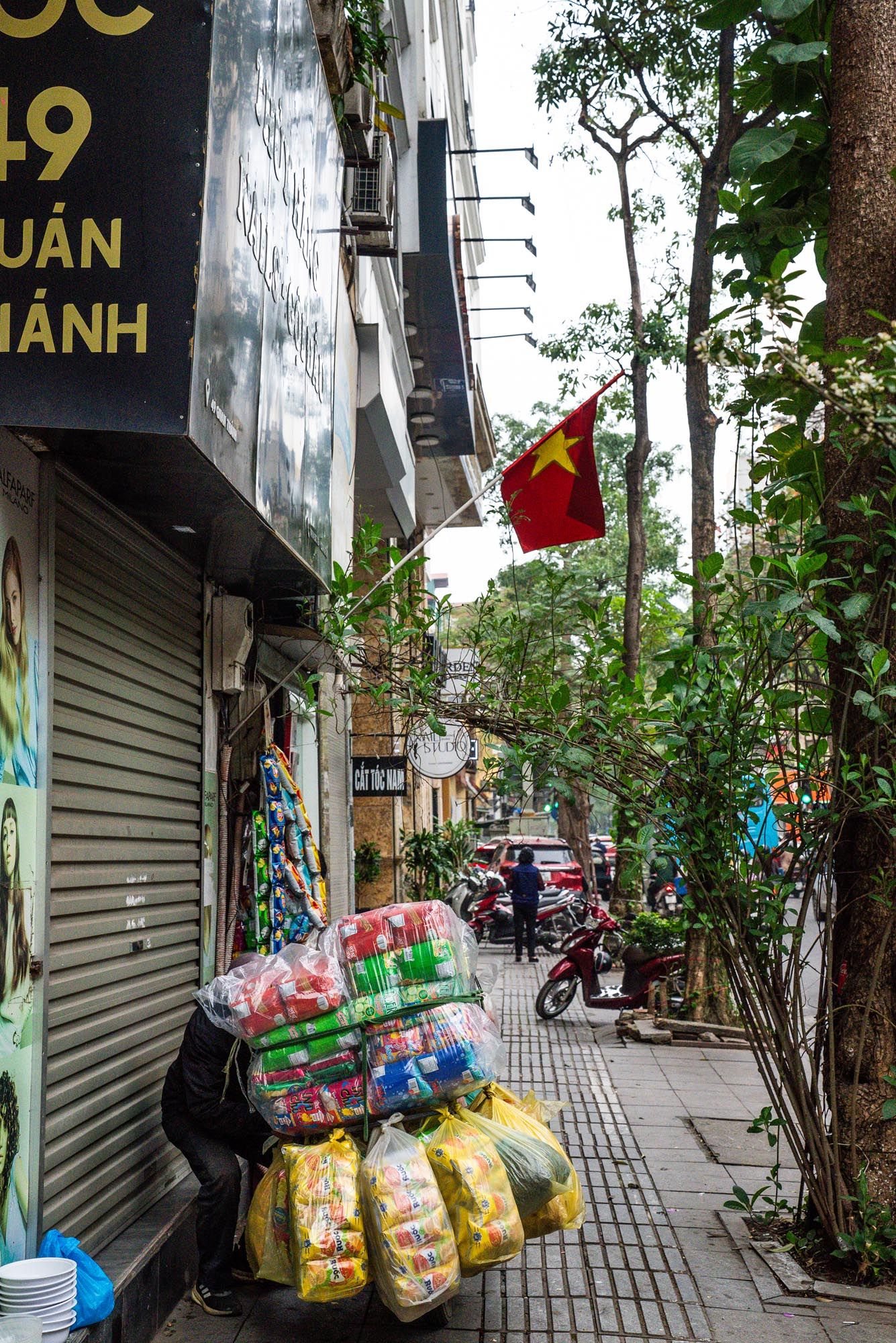 Bags of snacks and goods displayed outside closed storefronts on a quiet urban sidewalk in Vietnam.