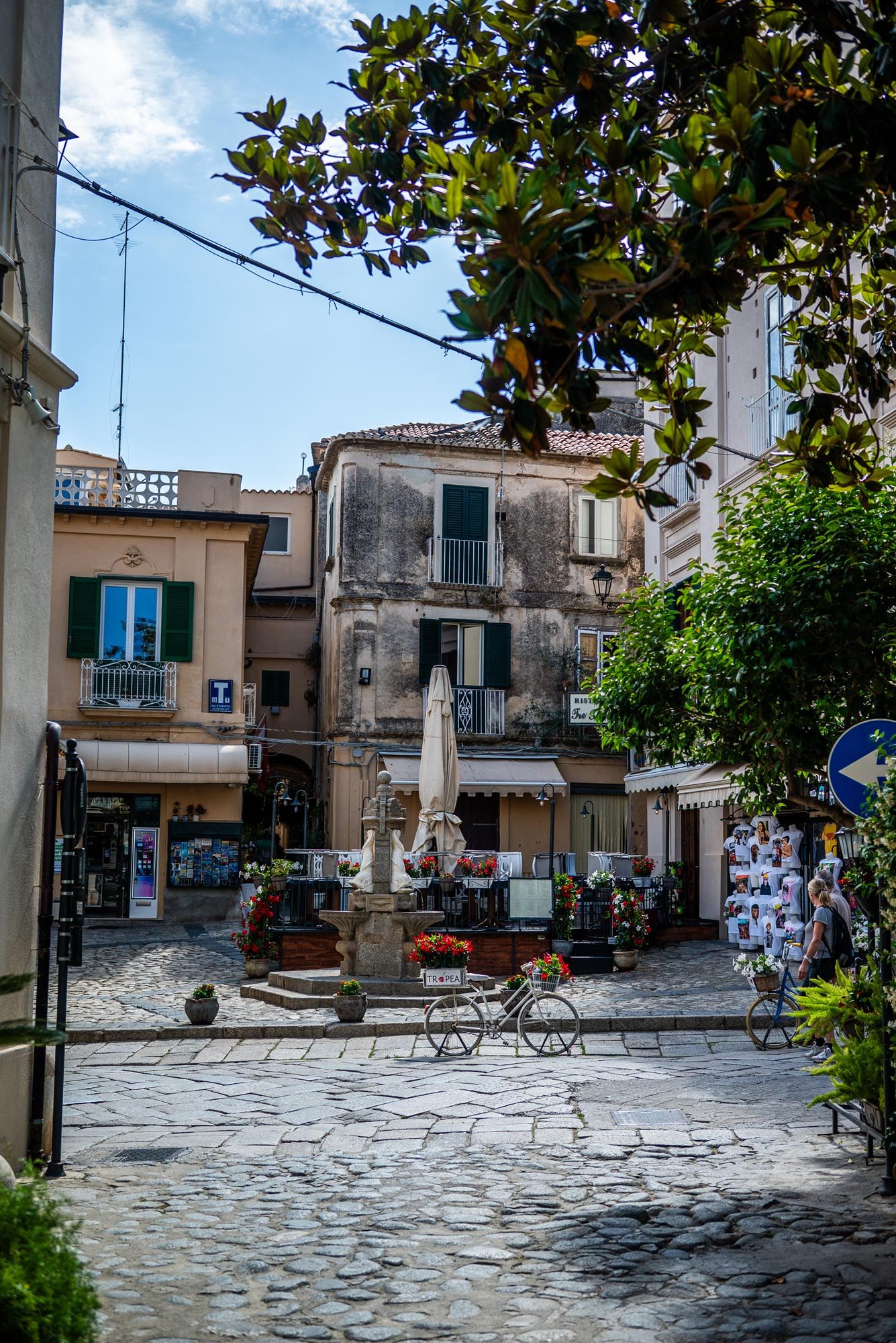 Charming cobblestone square in an old Mediterranean town with rustic buildings, outdoor cafe seating and leafy foliage overhead.