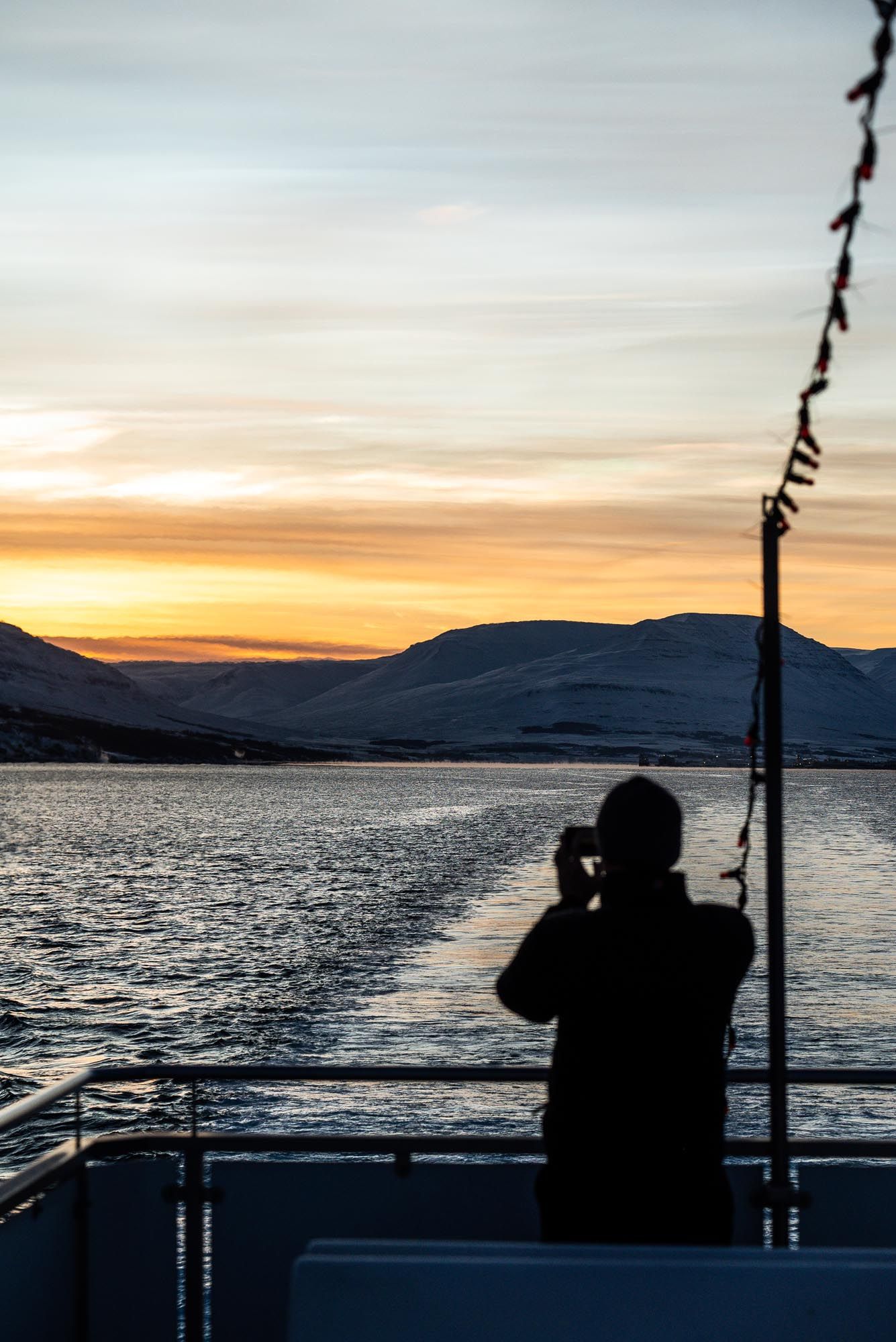 Silhouetted figure photographs sunset over mountain lake from boat deck during golden hour.