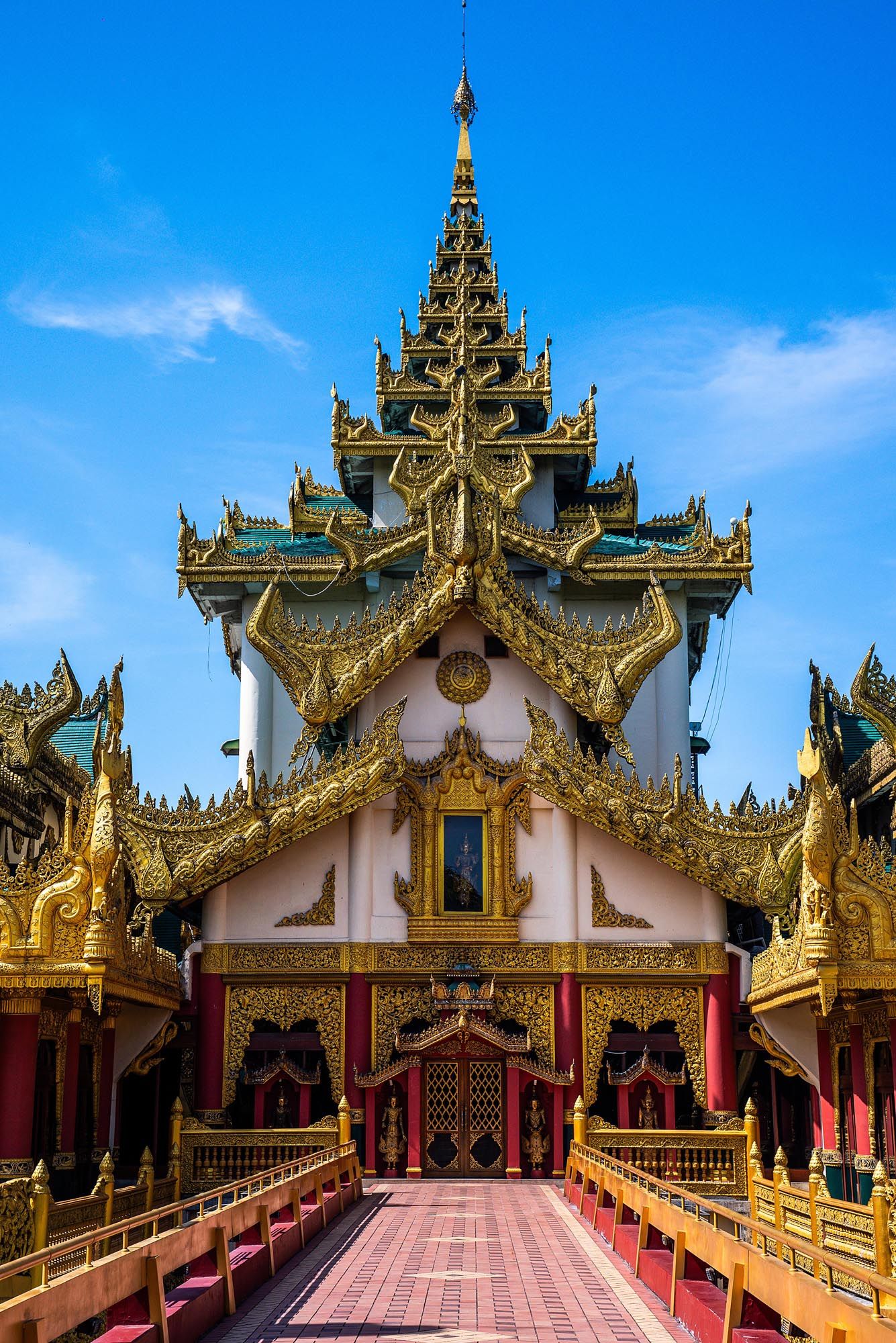 Ornate golden spires and decorative rooflines of a traditional Burmese Buddhist temple against a bright blue sky.