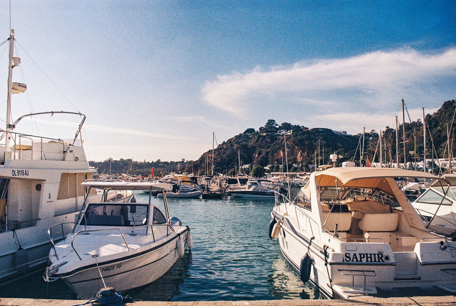 Luxury boats and yachts docked in a Mediterranean marina with hills in the background on a sunny day.