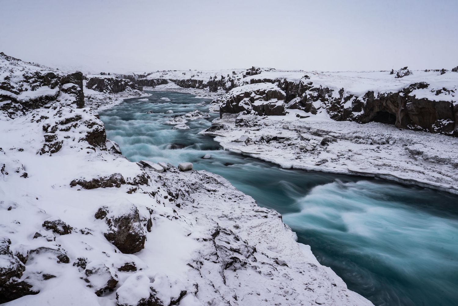 A turquoise river flows through snowy rocky terrain in Iceland during winter, creating a dramatic landscape.