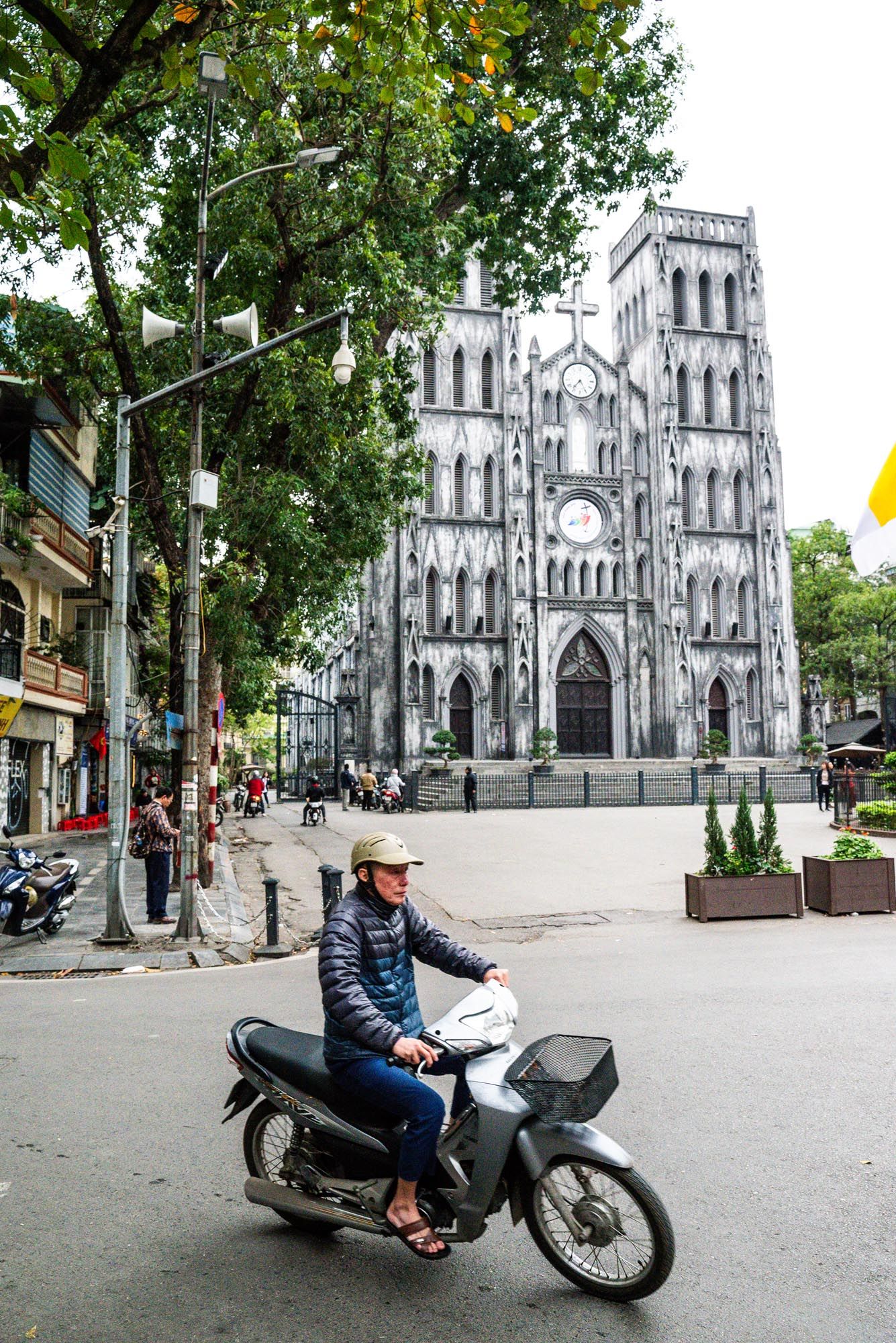 A motorcyclist rides past the Gothic St. Joseph's Cathedral in Hanoi, Vietnam with its twin spires and ornate facade.