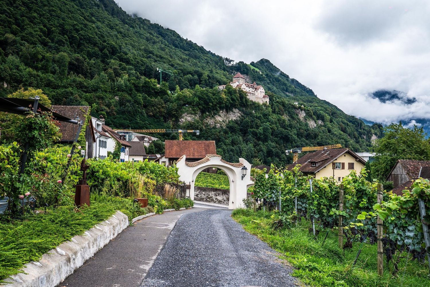 Winding mountain road through lush green hillsides with scattered houses and cloudy skies in a rural village setting.