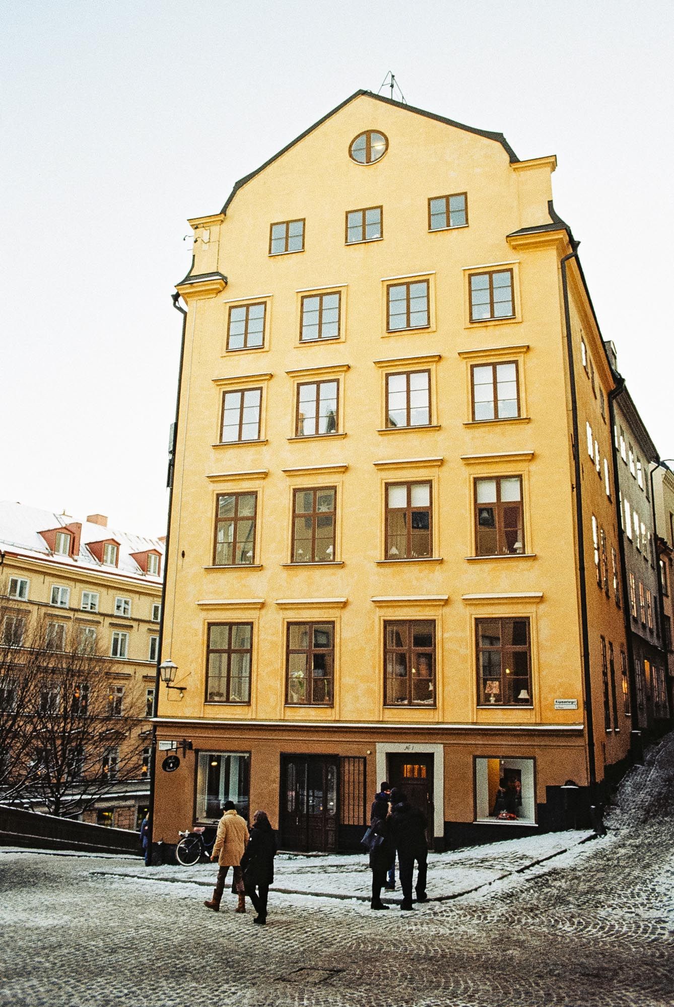 Historic yellow apartment building with multiple floors stands on a cobblestone street corner in a European city setting.