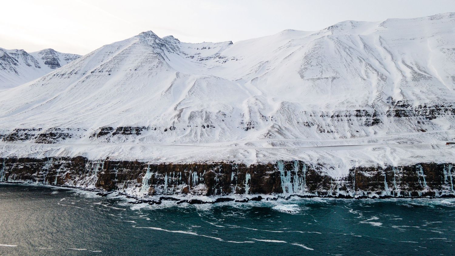 Dramatic cliffs and waterfalls along an icy coastline with snow-capped mountains in Iceland.