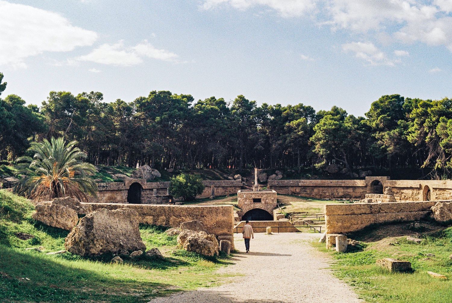 Ancient Roman amphitheater ruins with stone arches and pathways surrounded by trees and greenery in a park setting.