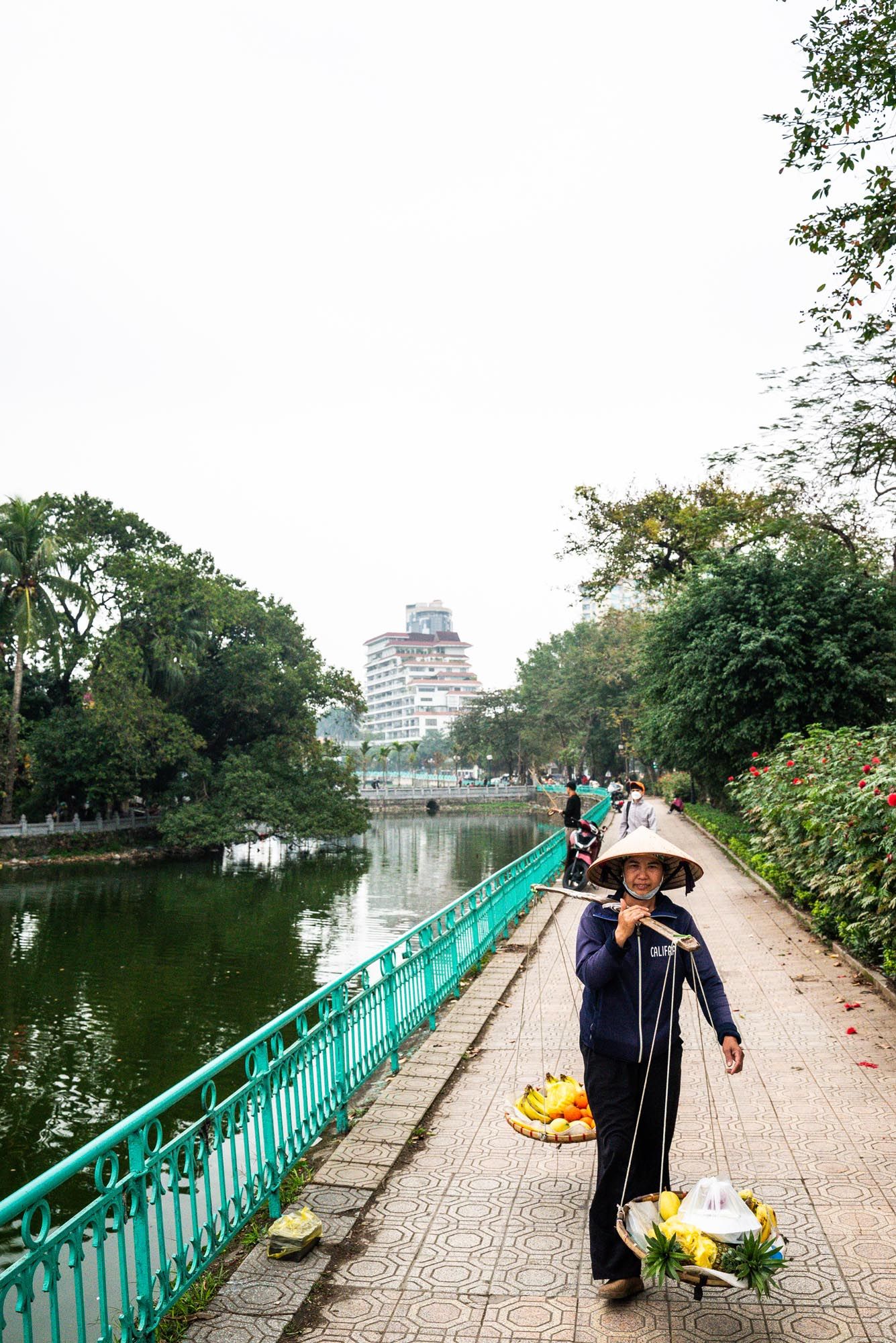 Flower vendor walking along a turquoise-railed pathway beside a tranquil pond in a Vietnamese city park.