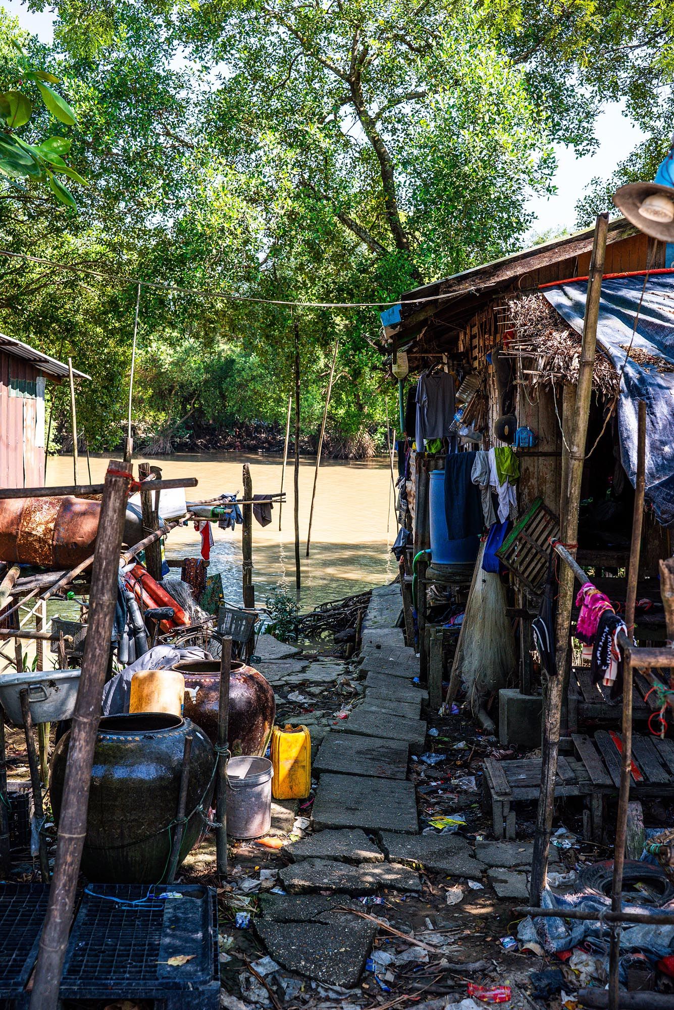 Makeshift shelters and scattered belongings line a dirt path in an outdoor homeless encampment under shady trees.