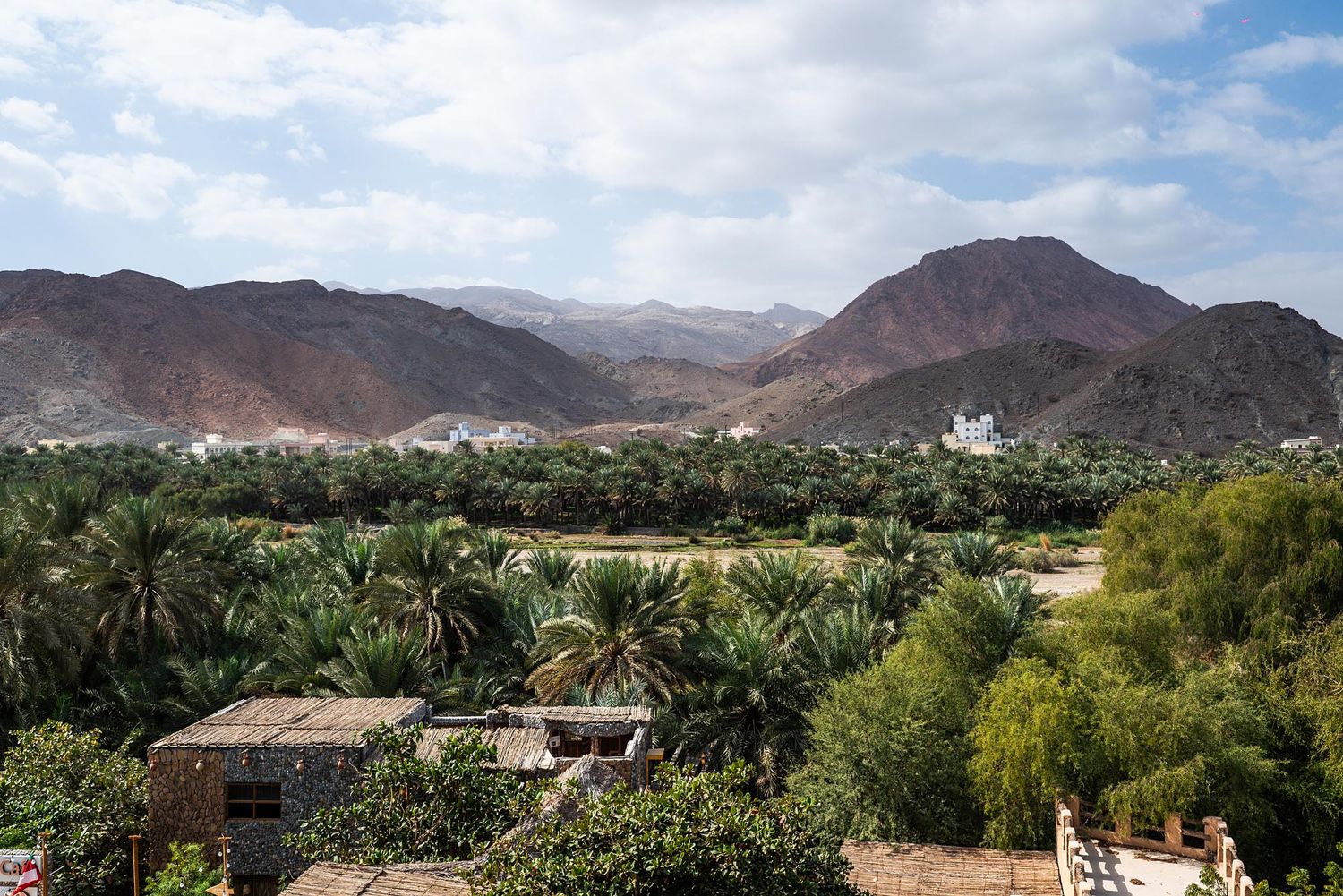 Panoramic view of rugged mountain ranges and desert landscape with scattered palm trees and buildings in the distance.