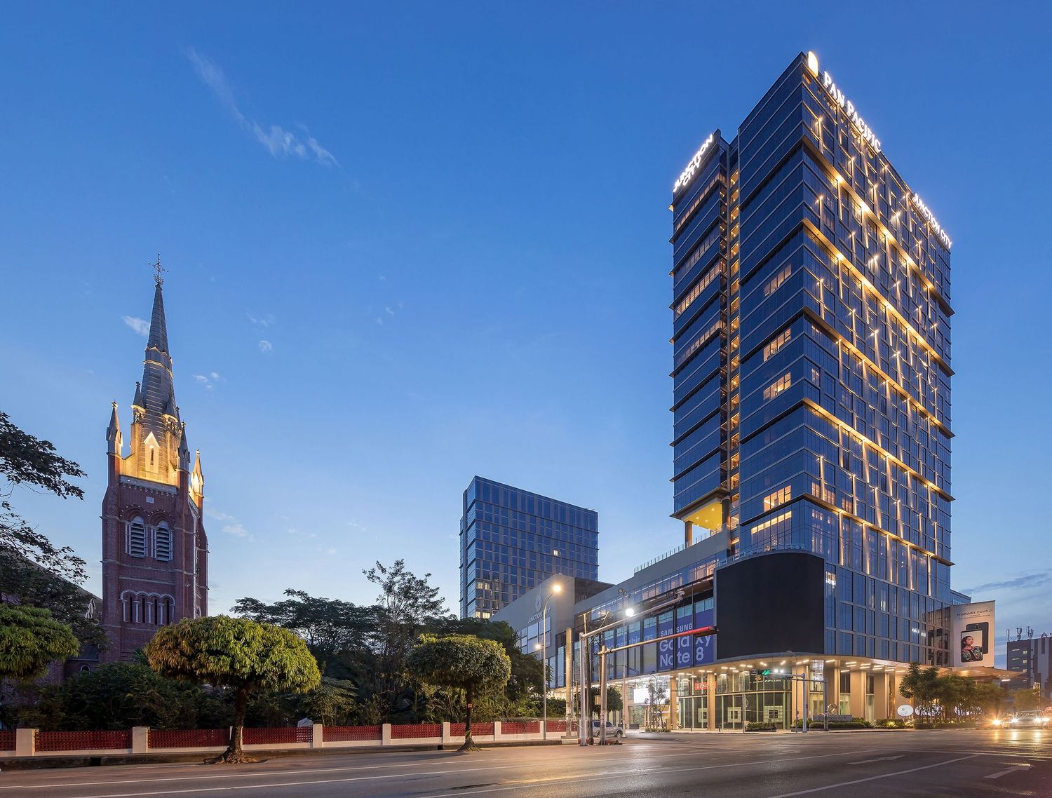 Modern glass office building with illuminated windows stands next to historic church spire against blue evening sky.