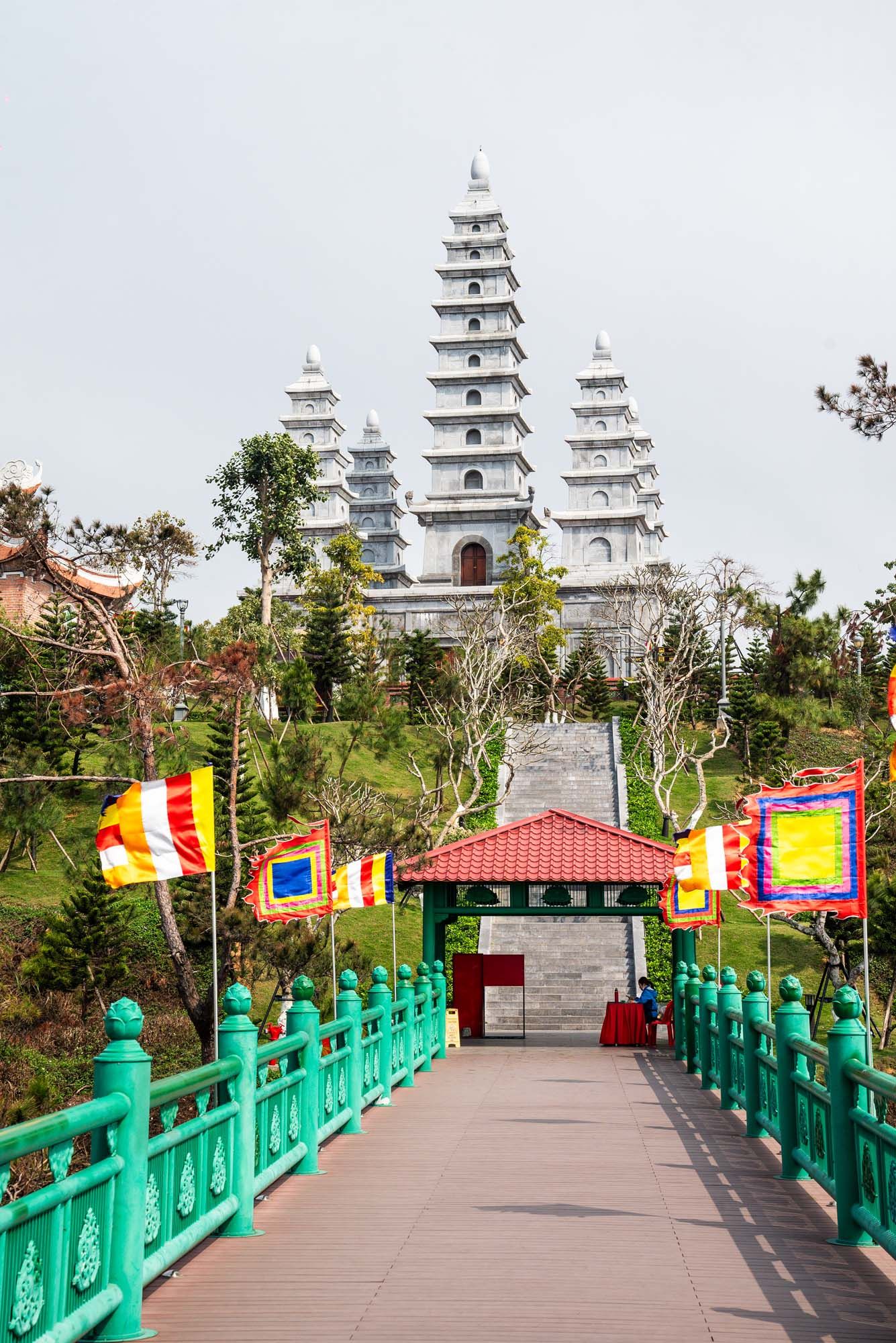 A panoramic view of a Buddhist temple entrance with turquoise railings, colorful flags, and white pagodas in the background.
