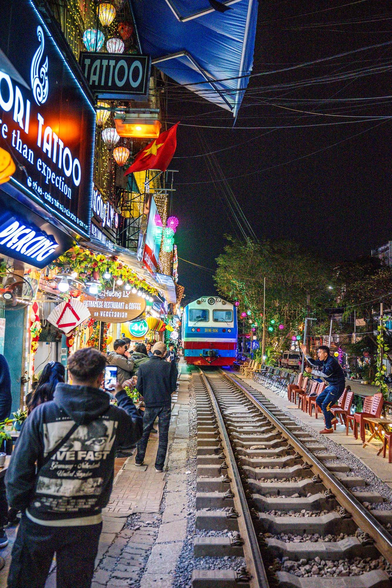 Illuminated train tracks run through a bustling night market street in Hanoi with colorful signs and shops along both sides.