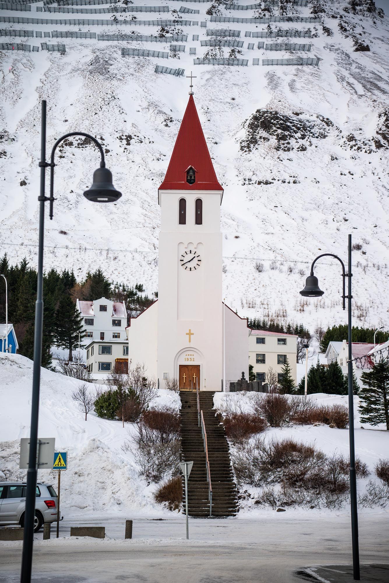 A small white church with a red spire stands against a snowy winter landscape in a mountain village.