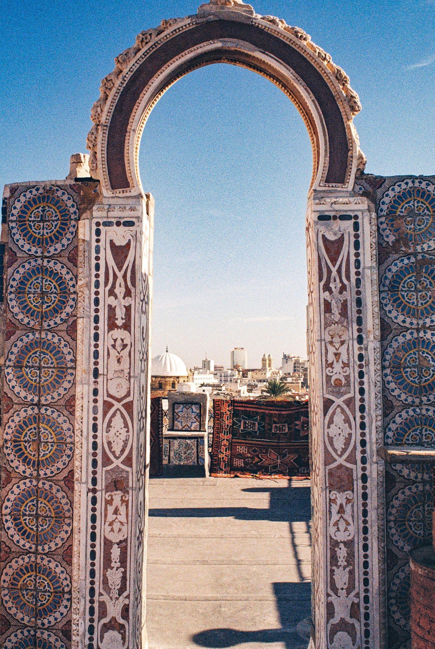 Ornate archway with intricate mosaic patterns and geometric designs frames a view of a traditional Middle Eastern cityscape.