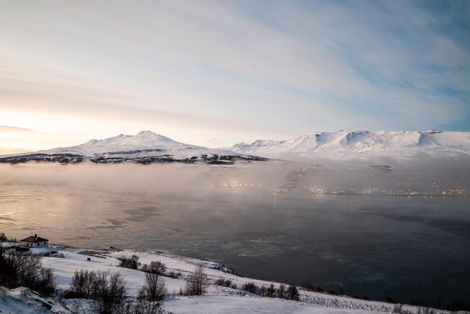 A panoramic winter landscape with snow-covered mountains and a misty fjord reflecting the cloudy sky in Norway.