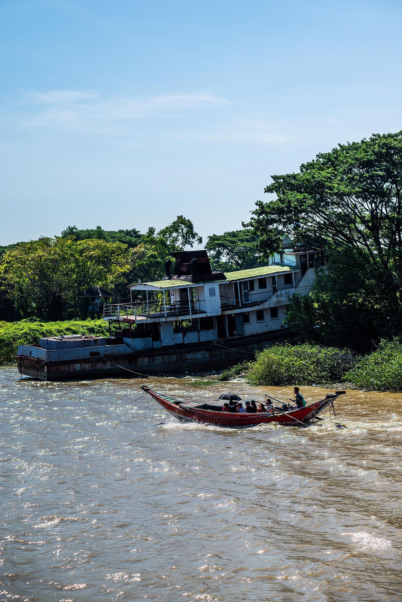 A large wooden riverboat navigates choppy waters near a forested shoreline on a sunny day.