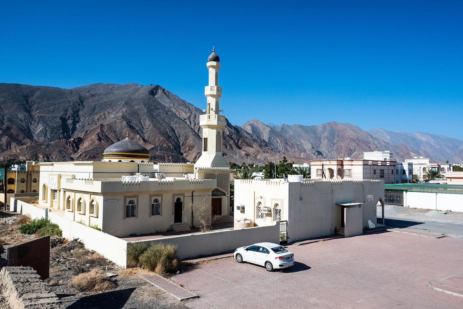 A white mosque with minaret stands against rugged mountain backdrop and bright blue sky in a desert landscape.