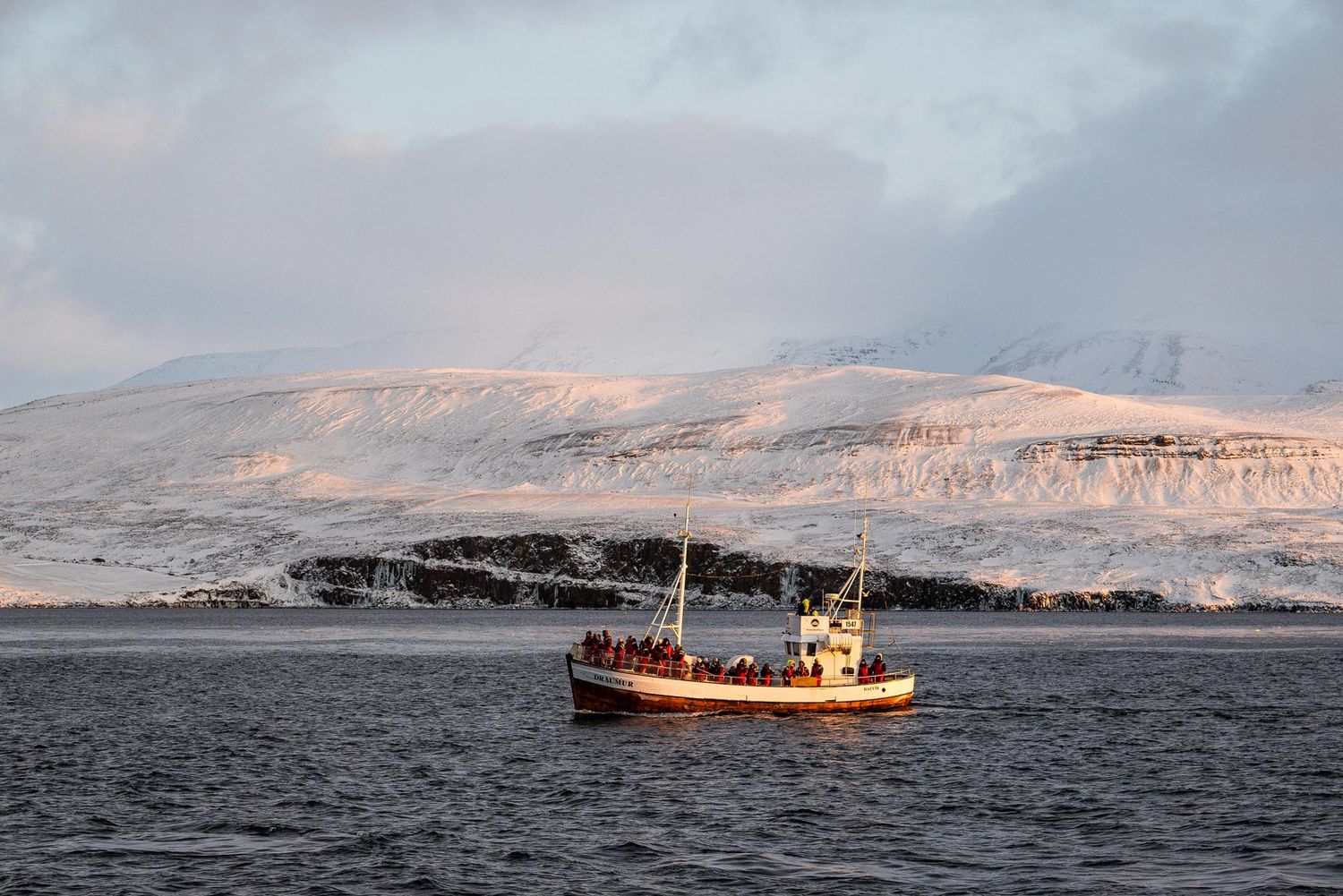 A fishing boat navigates through icy waters near snow-covered mountains in a dramatic winter seascape.