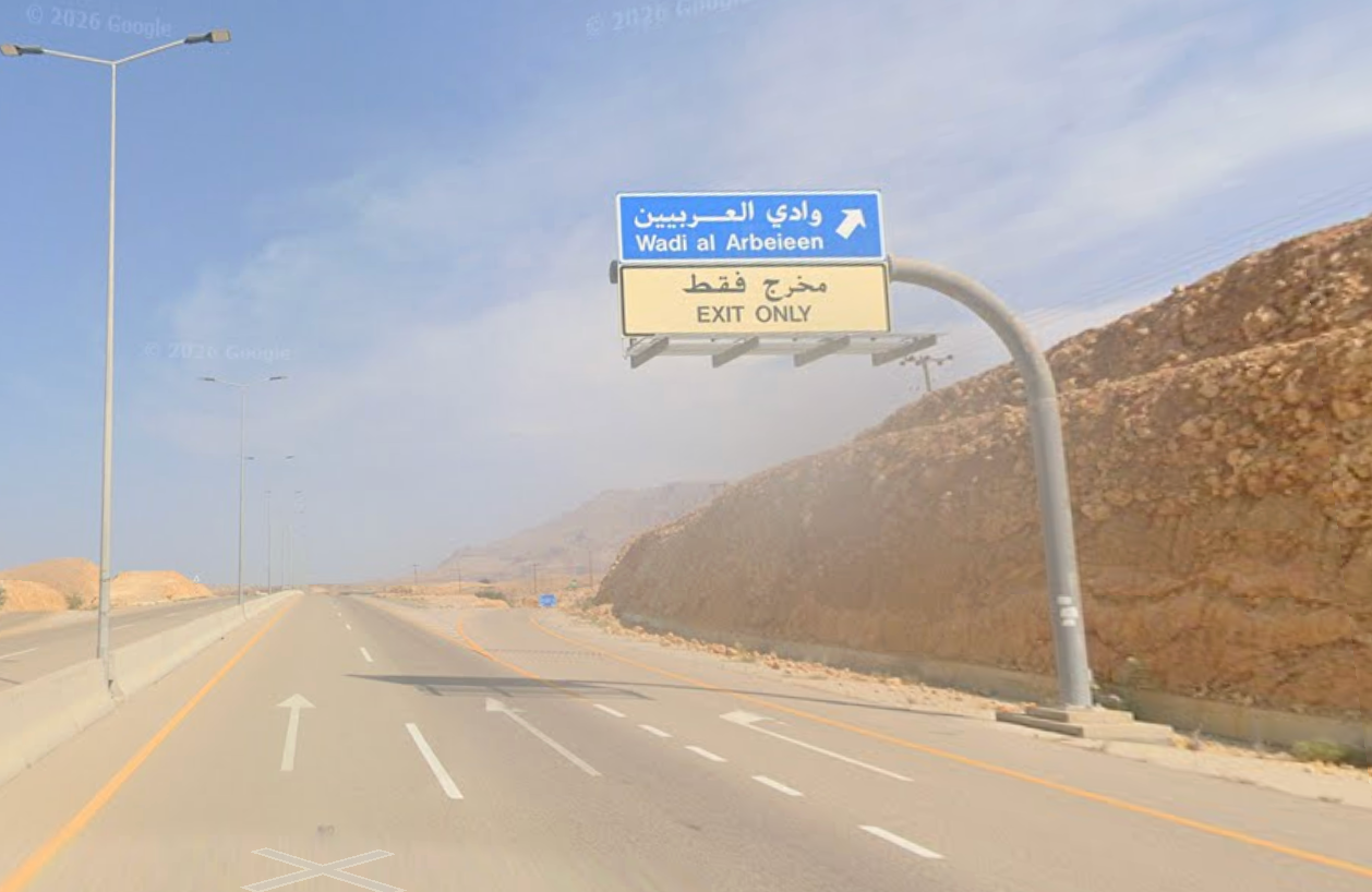 Desert highway curves through barren mountains with road signs and streetlights under a hazy blue sky.