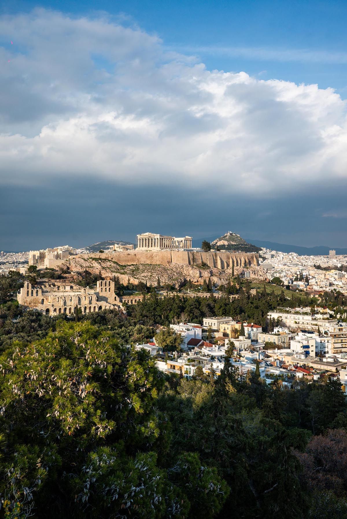 Panoramic view of Athens cityscape with the iconic Acropolis and Parthenon temple perched on the hilltop against a cloudy sky.