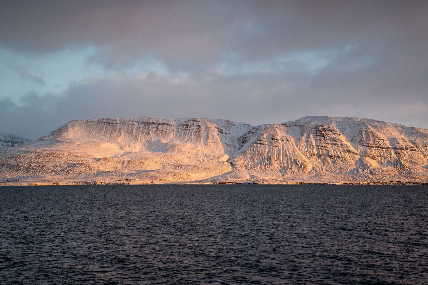 Dramatic sunset illuminates snow-dusted mountain cliffs rising above dark arctic waters in remote Nordic landscape.