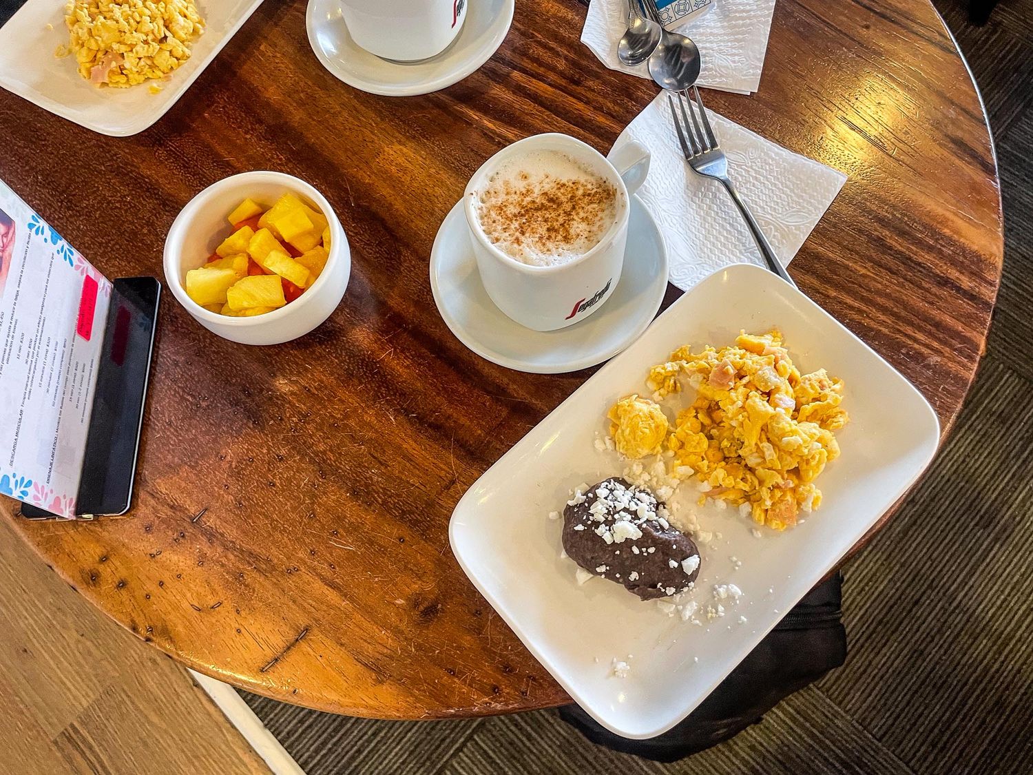 A breakfast spread on a wooden table with scrambled eggs, fruit, coffee, and pastries served on white plates.