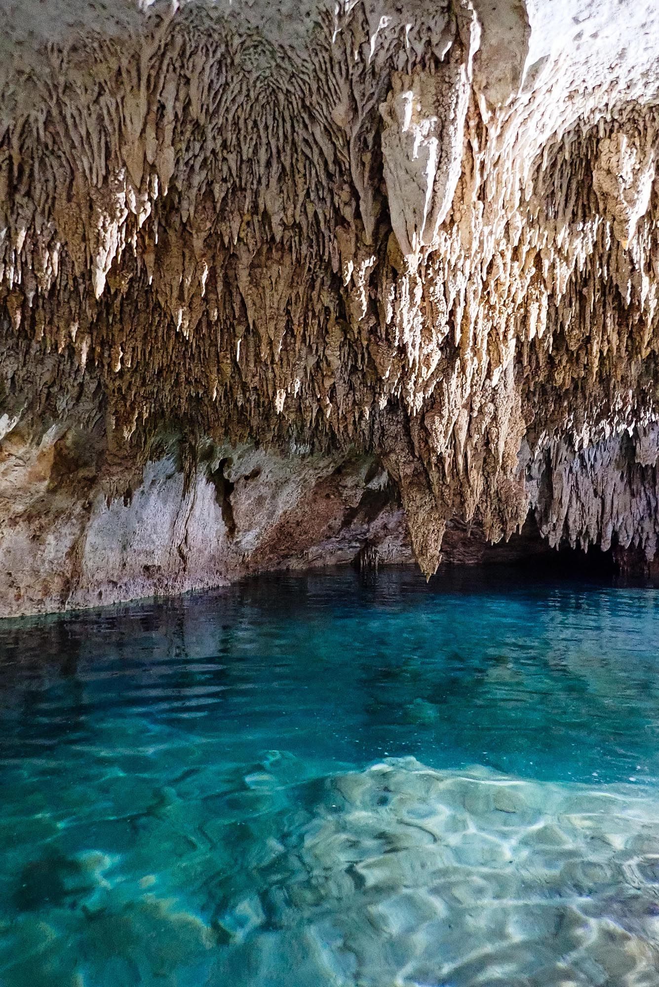 Stunning blue crystal-clear water reflects light beneath stalactites hanging from limestone cave ceiling.