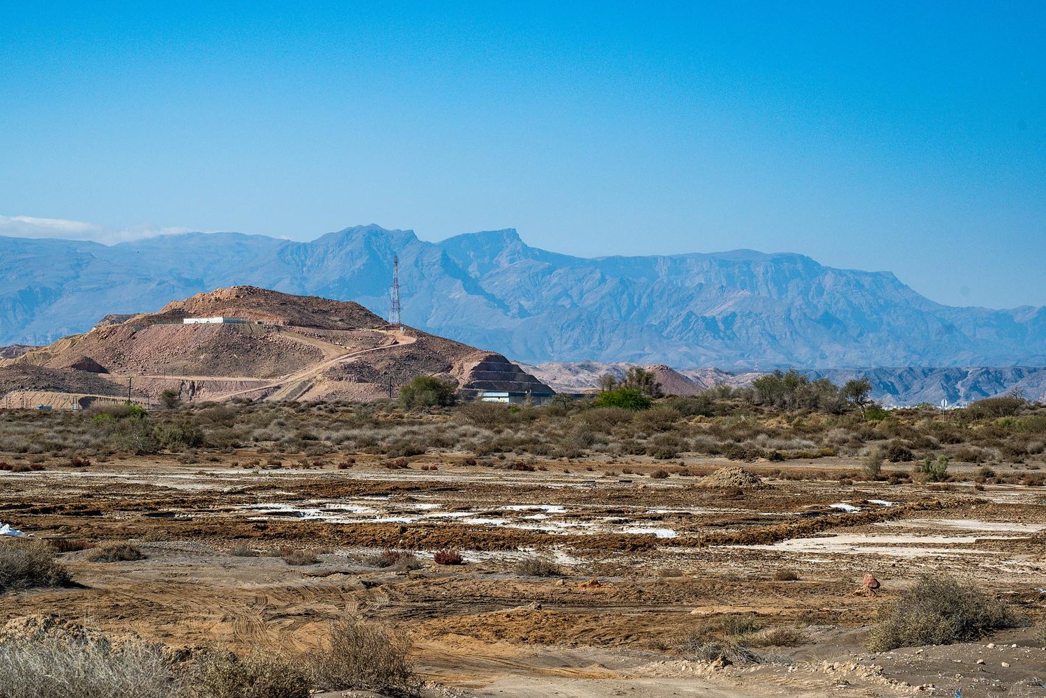 Rugged desert landscape with mountains in the background and a small structure on a hilltop under bright blue skies.
