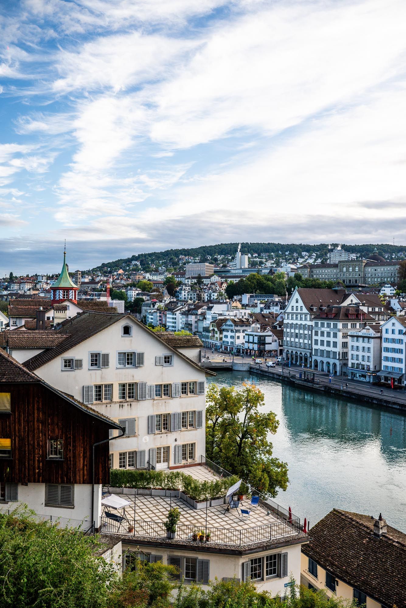Panoramic cityscape view of Zurich's historic waterfront buildings and Limmat River under cloudy blue skies.
