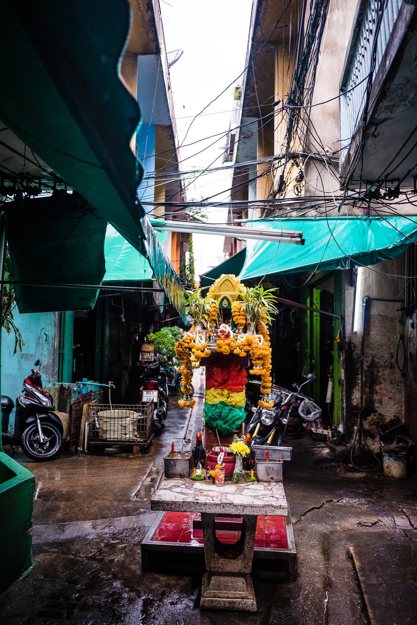 A small Buddhist shrine with yellow decorations sits in a narrow alleyway between turquoise-colored walls.