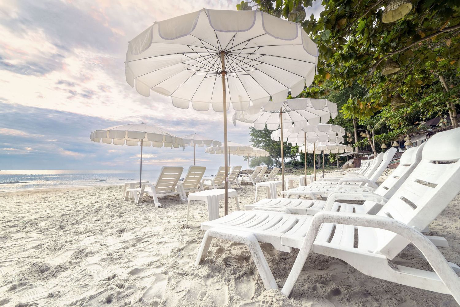 White beach loungers and umbrellas line a tropical shoreline with crystal clear turquoise waters and palm trees.