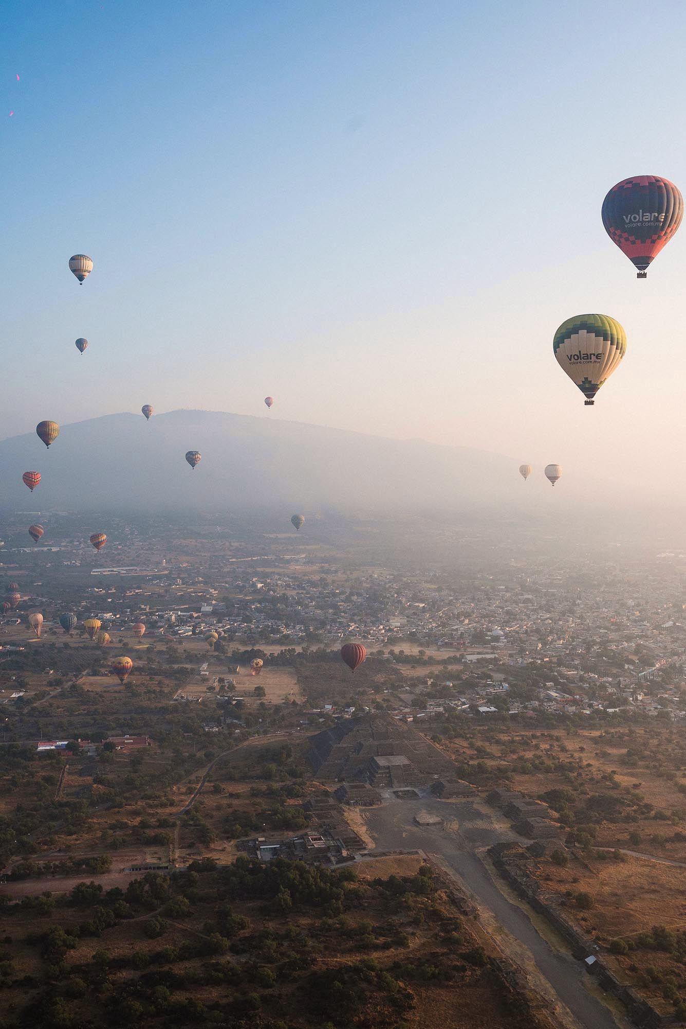 Hot air balloons float over a misty cityscape at dawn, creating a dreamy aerial panorama of urban and rural landscapes.
