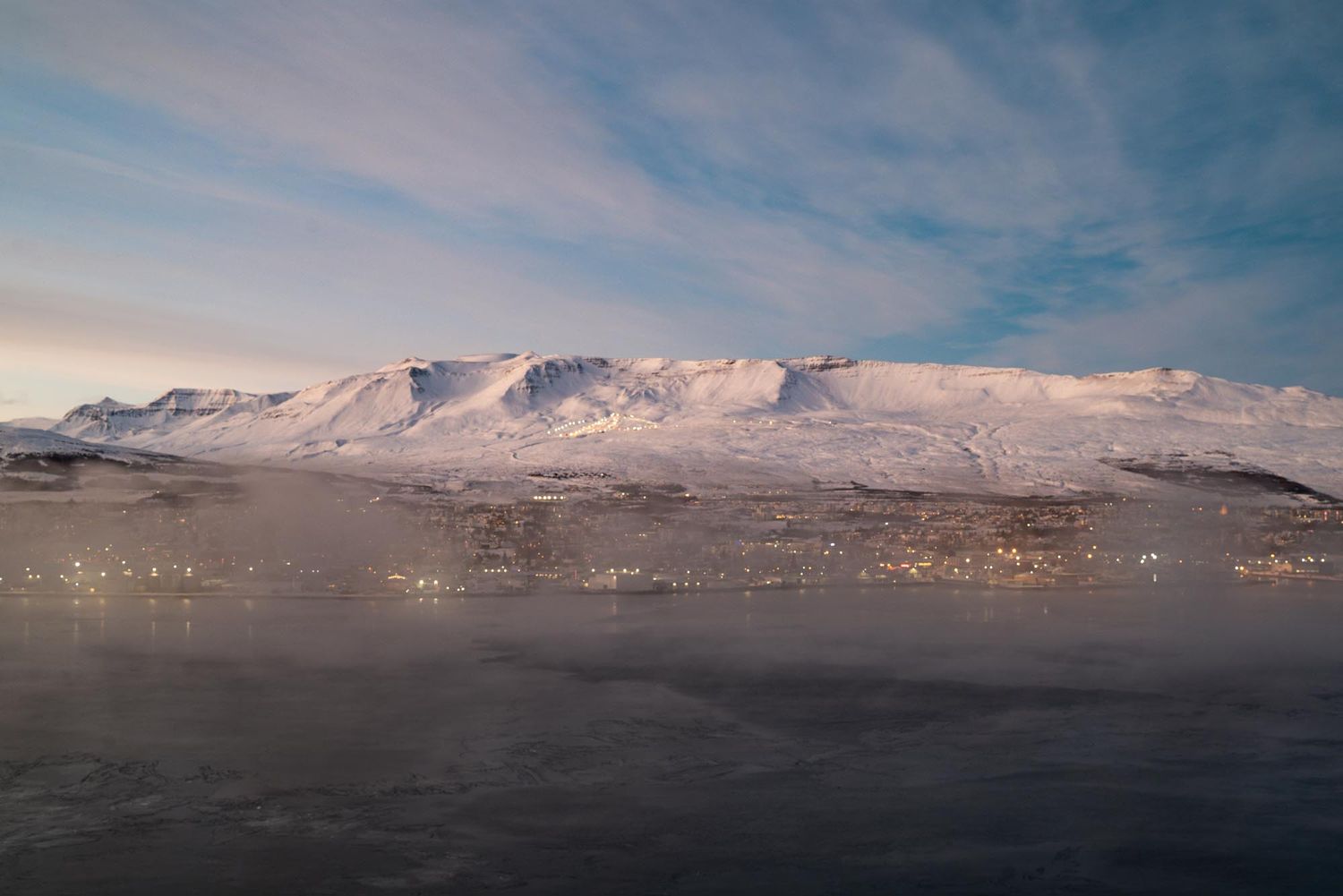 Snow-capped mountain range rises above misty waters under a pastel sky during golden hour.