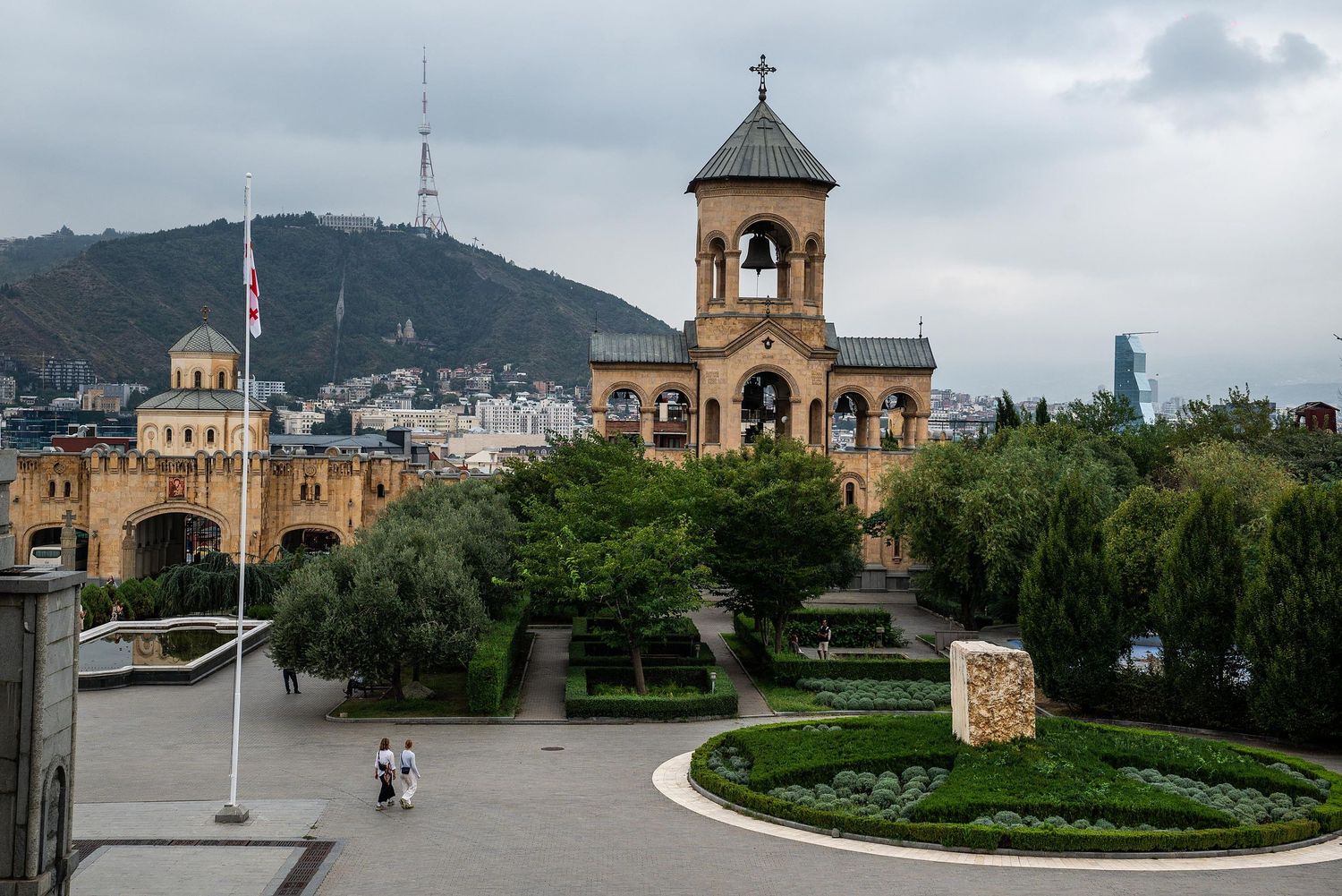 A panoramic view of Tbilisi city with historic churches and buildings nestled among green hills and cloudy skies.