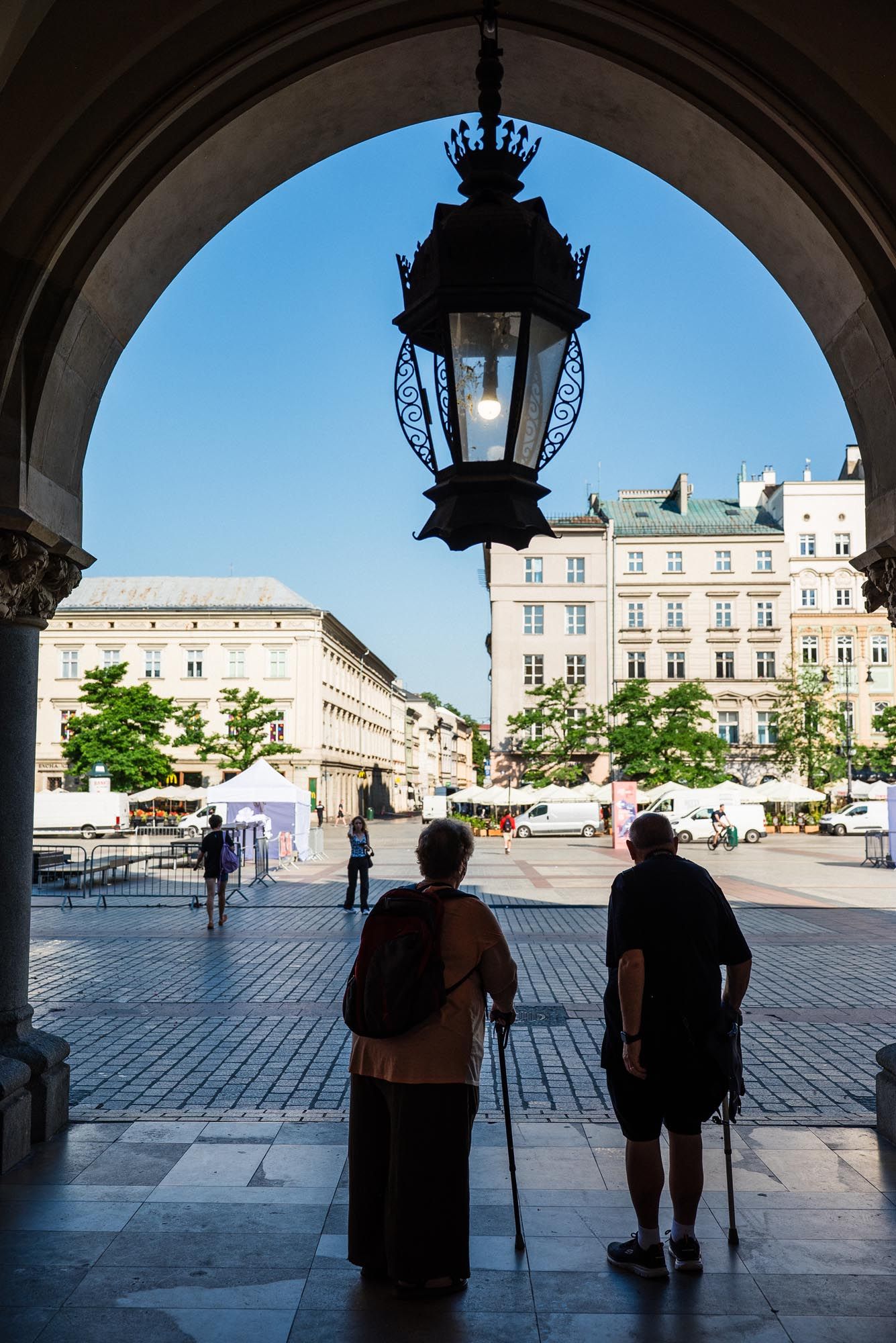 View through an archway of Krakow's Market Square with ornate hanging lamp and historic buildings on sunny day.