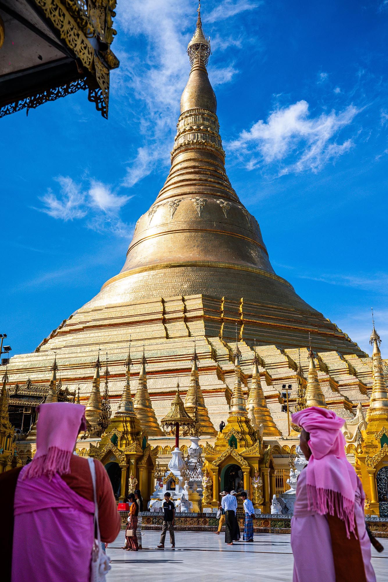 Golden stupa of Shwedagon Pagoda rises against blue sky with Buddhist statues and pink-robed monks in Yangon, Myanmar.